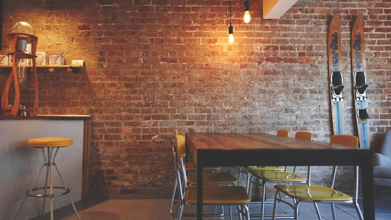 Interior of an empty coffee shop with a wooden table, yellow chairs and an exposed brick wall; no people visible.