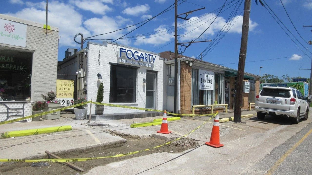 A suburban street with orange traffic cones and yellow caution tape around a torn-up sidewalk construction area; no people visible