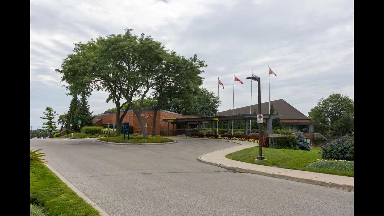 The brick Whitby Town Hall civic building on Rossland Road in Whitby, Ontario, photographed on an overcast day with trees, flagpoles, and a curved driveway in front; no people present.
