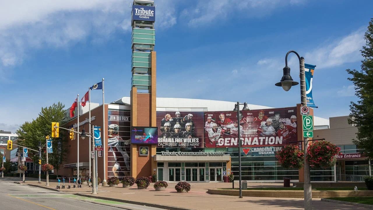 Exterior of the Tribute Communities Centre, home arena of the Oshawa Generals, on Athol Street in downtown Oshawa on a clear day; the brick facade carries large Oshawa Generals 'Generations' and Oshawa FireWolves mural wraps, team flags fly out front, and the arena's tall tower sign rises above the entrance.